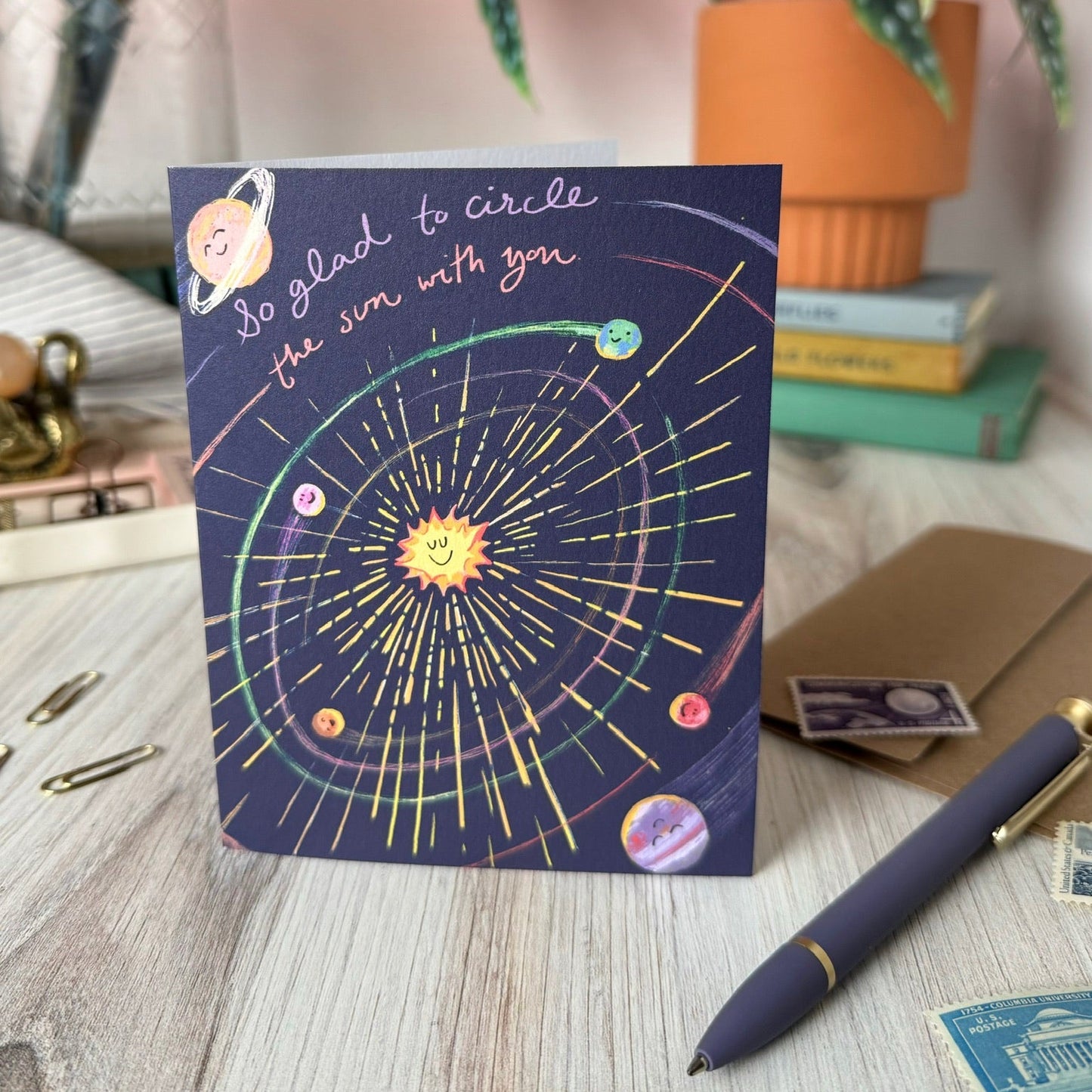 A greeting card featuring a colorful solar system illustration on a dark navy background, styled on a light wood desk with a pen, envelope, and books in the background. The card says "so glad to circle the sun with you."