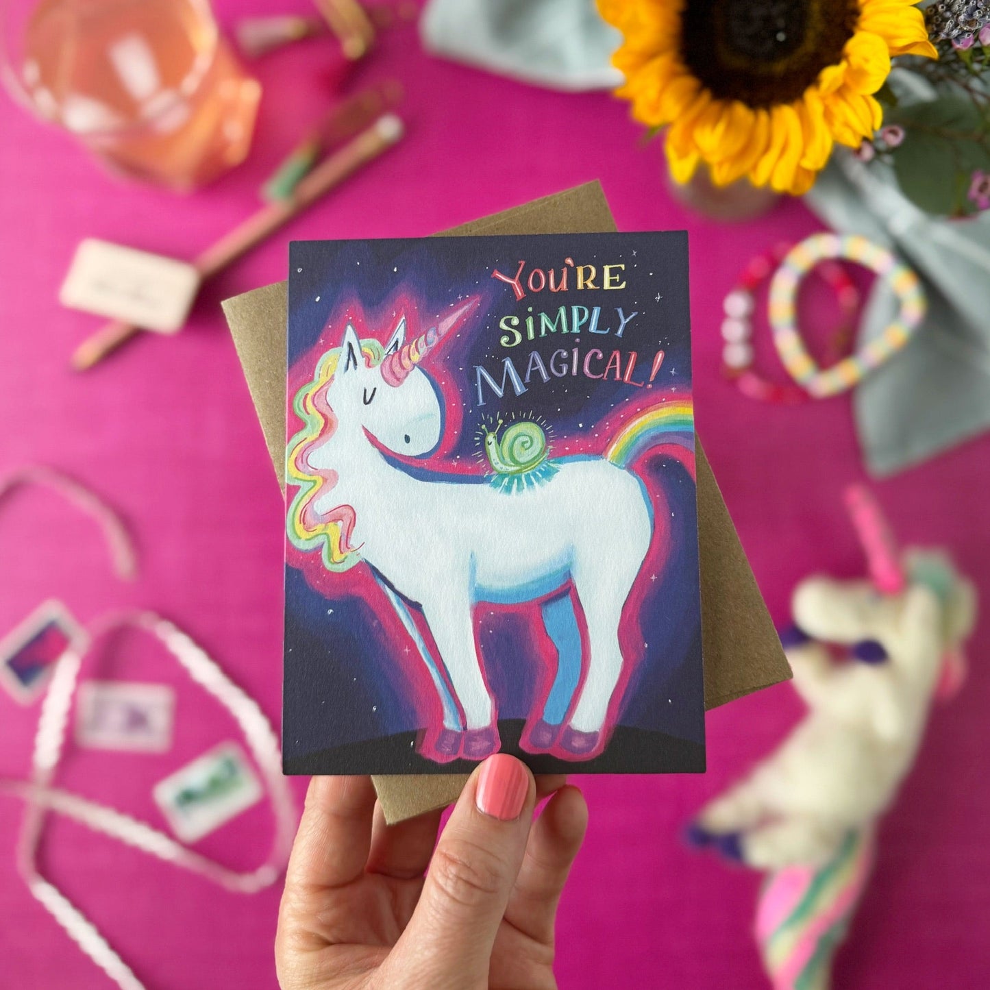 A hand holds a square greeting card featuring a colorful unicorn and snail illustration with the text 'YOU'RE SIMPLY MAGICAL!'. The card is set against a bright pink styled flat-lay background with a sunflower, ribbon, and other decorative items.