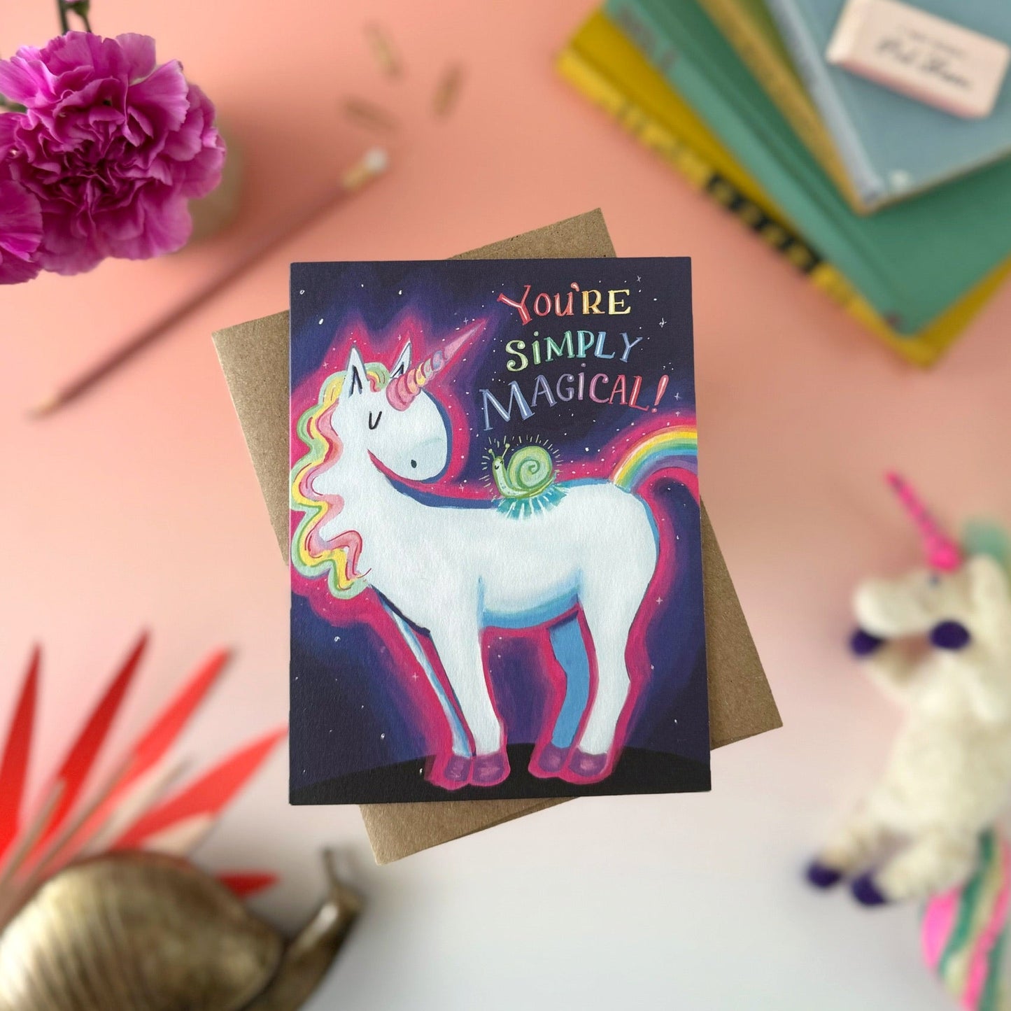 A square greeting card featuring a colorful unicorn and snail illustration with the text 'You're Simply Magical!' on a dark starry background, styled on a kraft envelope with flowers, books, and a plush toy around it.