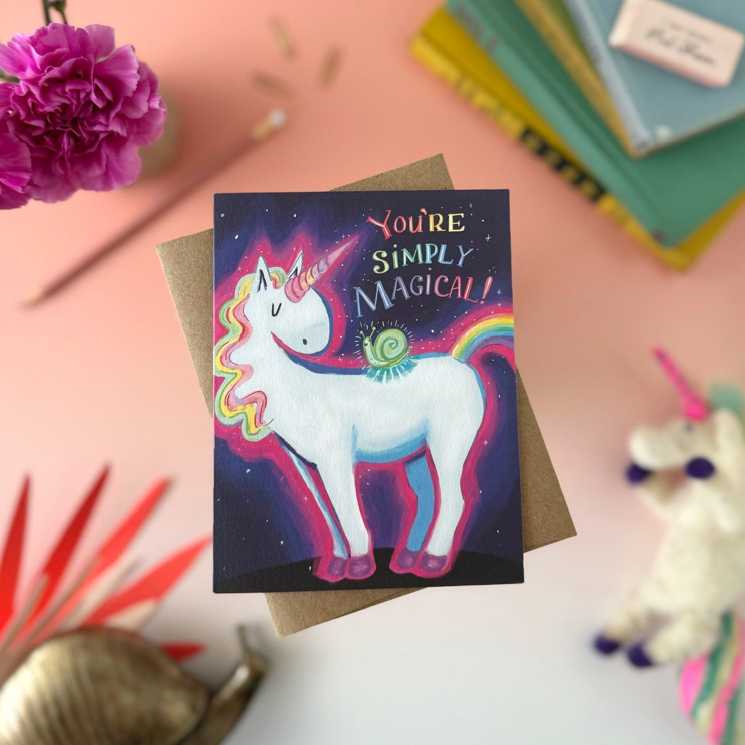 A square greeting card featuring a colorful unicorn and snail illustration with the text 'You're Simply Magical!' on a dark starry background, styled on a kraft envelope with flowers, books, and a plush toy around it.
