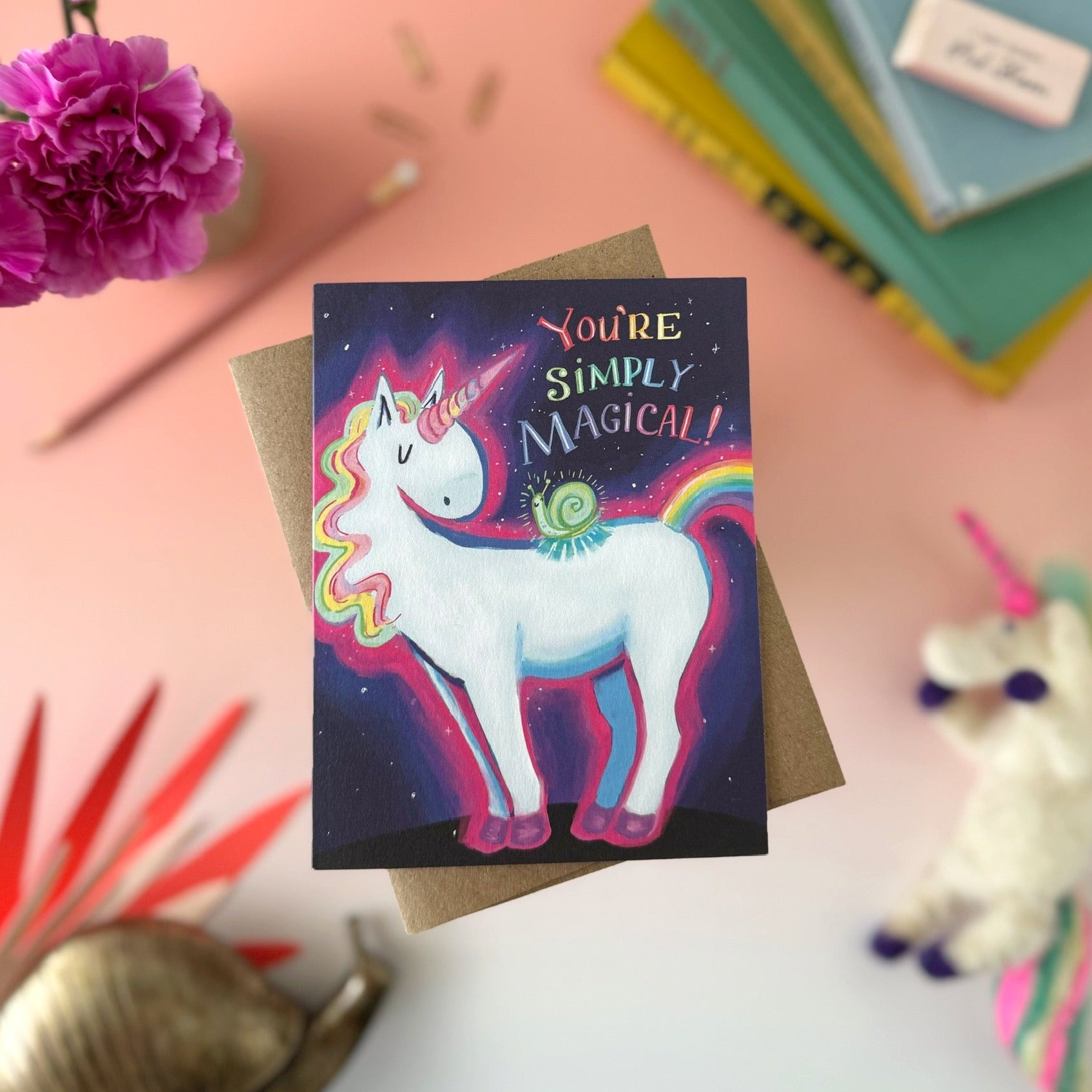 A square greeting card featuring a colorful unicorn and snail illustration with the text 'You're Simply Magical!' on a dark starry background, styled on a kraft envelope with flowers, books, and a plush toy around it.