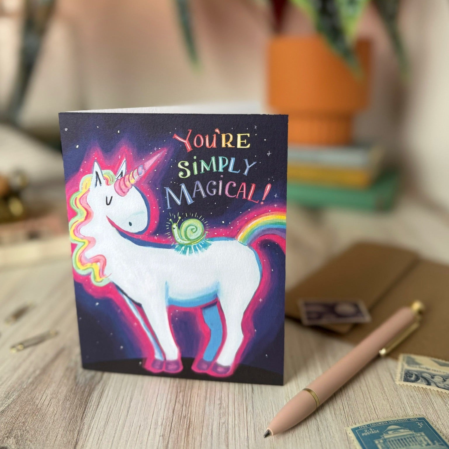 A square greeting card featuring a colorful unicorn illustration with the text 'YOU'RE SIMPLY MAGICAL!' stands on a light wood tabletop. The card is photographed in a styled indoor setting with a blurred plant, books, envelope, pen, and postage stamps in the background.